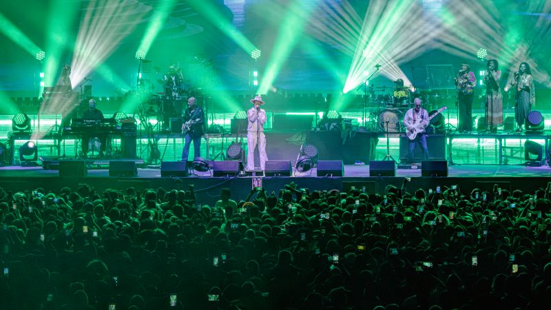 Ambiente lleno en la Arena CDMX para Jamiroquai Vista frontal del escenario bañada en luces verdes con la alineación completa de Jamiroquai y la multitud con celulares encendidos.