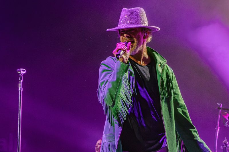 Jay Kay con sombrero brillante en el clímax del show Primer plano de Jay Kay cantando con un sombrero brillante plateado y su chaqueta verde sobre fondo púrpura.