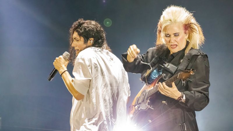 Jennifer Batten y el intérprete en This Is Michael El intérprete del tributo cantando en un micrófono blanco junto a la guitarrista Jennifer Batten tocando su guitarra eléctrica iluminados por luces de escenario.