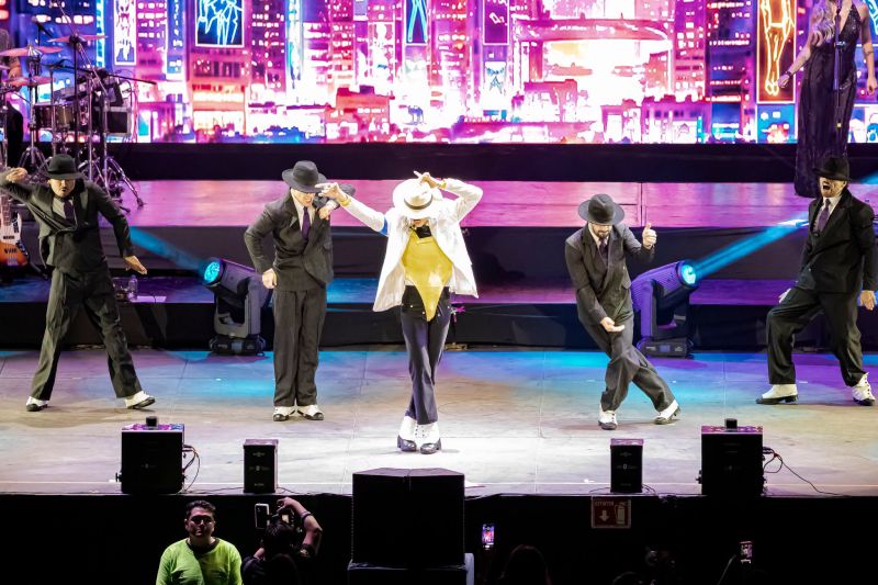 Coreografía de Smooth Criminal en la Arena CDMX El intérprete luciendo traje blanco, sombrero y leotardo dorado ejecutando la clásica inclinación de Smooth Criminal junto a sus bailarines vestidos de traje oscuro en el escenario.
