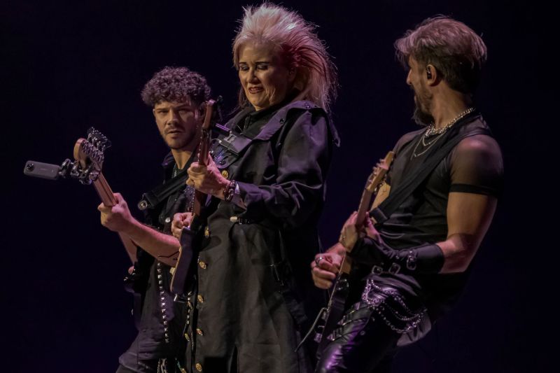 Duelo de guitarras con Jennifer Batten Jennifer Batten tocando la guitarra al centro flanqueada por el bajista y otro guitarrista del grupo musical sobre un fondo oscuro.