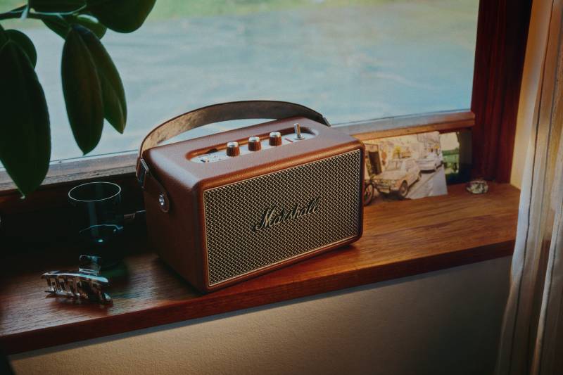 Bocina portátil en ambiente cálido Dispositivo de audio con diseño clásico colocado sobre una repisa de madera, acompañado por objetos decorativos y una iluminación suave que resalta su estética retro.