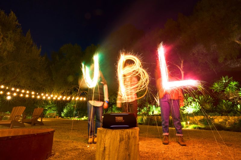 JBL – Nueva generación portátil Tres personas sostienen luces de bengala que forman letras luminosas mientras una bocina JBL se muestra en primer plano sobre un tronco.