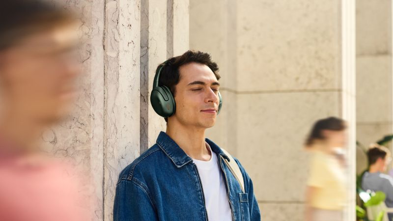 Persona con audífonos verdes de diadema disfrutando audio en un entorno urbano mientras permanece con los ojos cerrados.