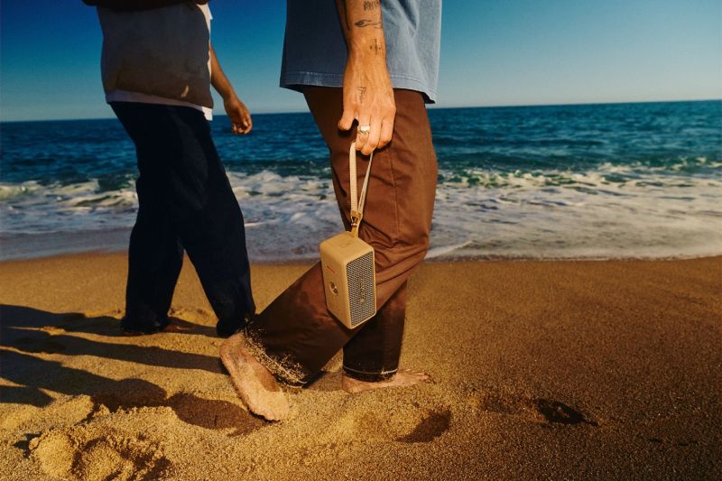 Bocina portátil Marshall Emberton III Brown en la playa Una persona caminando descalza por la playa sosteniendo la bocina portátil Marshall Emberton III Brown mediante su correa, con el mar de fondo bajo la luz del sol.
