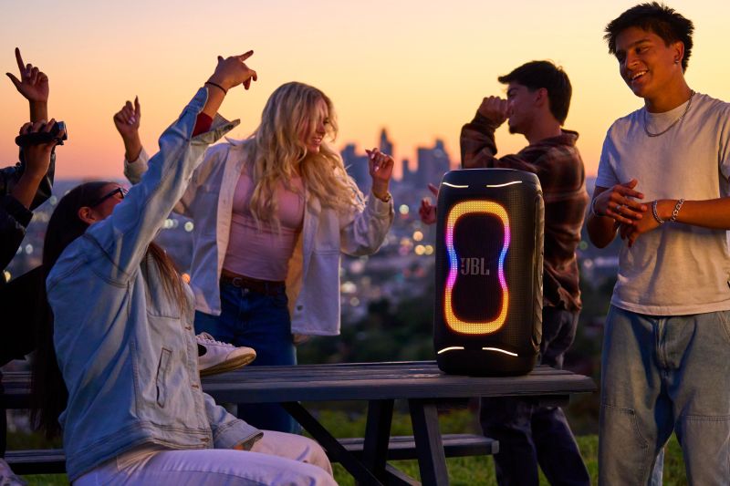 JBL PartyBox 130 en reunión al aire libre Grupo de jóvenes bailando y disfrutando al aire libre al atardecer en una terraza con vista a la ciudad, junto a una bocina portátil JBL PartyBox 130 encendida sobre una mesa de madera con luces LED dinámicas en tonos cálidos.