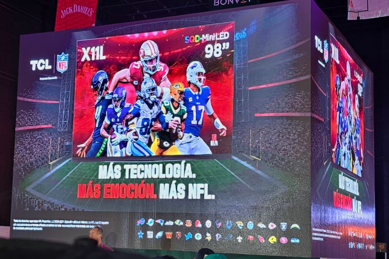 Presentación de la pantalla TCL X11L de 98 pulgadas con patrocinio de la NFL Una pantalla gigante cúbica en un evento de presentación donde se muestra la televisión TCL X11L de 98 pulgadas con tecnología SQD-MiniLED y gráficos de jugadores de la NFL sobre una cancha virtual.