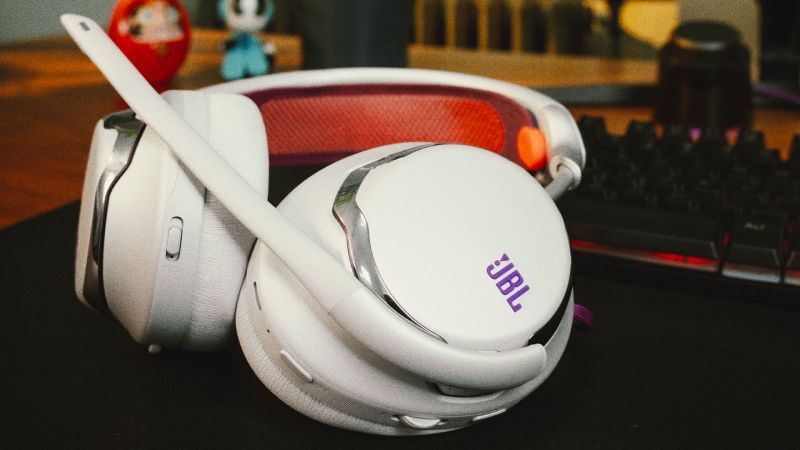 Audífonos para videojuegos JBL Quantum 950 de color blanco con detalles morados y naranja sobre un escritorio junto a un teclado gamer retroiluminado.