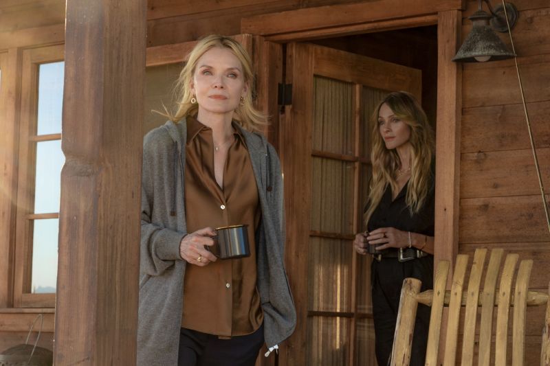 The Madison – Renovación temporada 3 Dos personas en la entrada de una cabaña de madera, una sosteniendo una taza y otra de pie en el umbral, en un entorno natural iluminado suavemente.