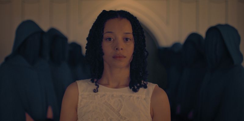Una persona con vestido blanco se sitúa en primer plano, mientras varias figuras encapuchadas y vestidas de oscuro se alinean simétricamente en el fondo. El espacio interior tiene arcos y una iluminación tenue.