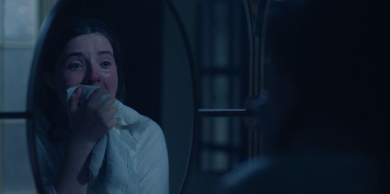 Una persona observa su reflejo en un espejo dentro de una habitación oscura. Sostiene un paño cerca del rostro, iluminada por una luz azulada que acentúa la atmósfera introspectiva.