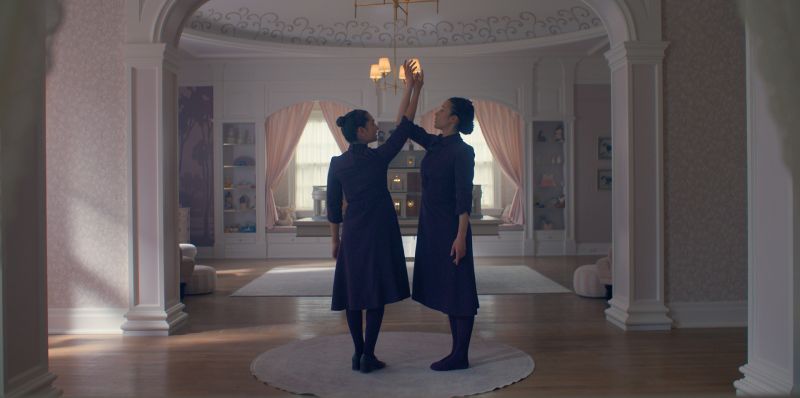Dos personas vestidas con atuendos oscuros se enfrentan en una sala elegante, levantando un brazo en un gesto simétrico. El espacio está decorado con arcos, cortinas claras y mobiliario ordenado.