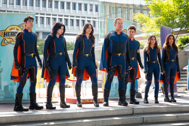Elenco principal vistiendo trajes de superheroes en la pelicula La Furia de los Thundermans de Paramount Plus y Nickelodeon Fotografía de estudio en plano entero del elenco principal de la película "La Furia de los Thundermans". De izquierda a derecha, siete miembros del reparto (Diego Velázquez, Ariel Winter, Kira Kosarin, Chris Tallman, Jack Griffo, Addison Riecke y Rosa Blasi) visten trajes tácticos ajustados de superhéroes en azul marino con detalles en naranja e insignias con la letra 'T' en el pecho y cinturones, posando alineados al aire libre frente a una pancarta del 'Hall of Heroes'.