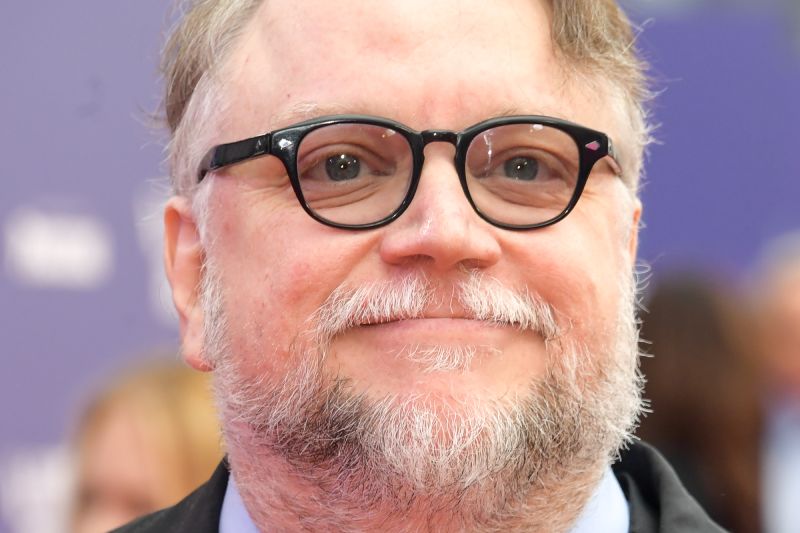 Guillermo del Toro ponente en el Crunchyroll Anime Future Forum 2026 Fotografía de retrato en plano medio del cineasta Guillermo del Toro sonriendo hacia la cámara. Viste una chaqueta oscura, camisa gris claro y lleva sus característicos lentes de armazón negro con barba y cabello canoso. El fondo presenta un tono azul violeta fuera de foco.