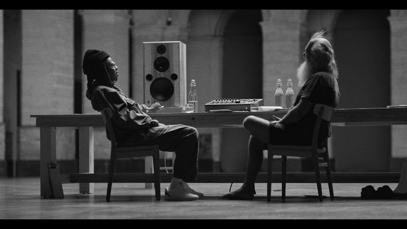“Jaÿ-Z in 8” se estrena en exclusiva por MUBI Fotografía en blanco y negro donde se aprecia a Jay-Z y al productor Rick Rubin sentados frente a frente en sillas de madera a los extremos de una mesa larga. Al centro de la mesa se observa un altavoz de estudio y equipo de sonido. Jay-Z, del lado izquierdo, luce vestimenta casual con gorro y calzado blanco, mientras Rick Rubin, del lado derecho, viste playera oscura, shorts y luce su característica barba larga y canosa.