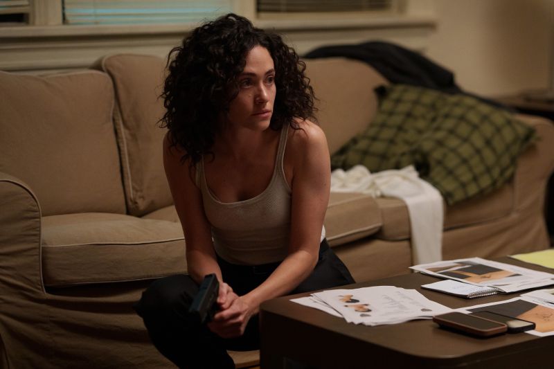 La actriz Emmy Rossum caracterizada como la agente del FBI Alice Black en la serie 'Furia'. Se encuentra agachada vestida con una playera de tirantes clara y pantalón oscuro, sosteniendo una pistola con la mano derecha frente a una mesa de centro que contiene expedientes, fotografías y teléfonos móviles. Al fondo se observa un sillón color beige con ropa y un cojín.