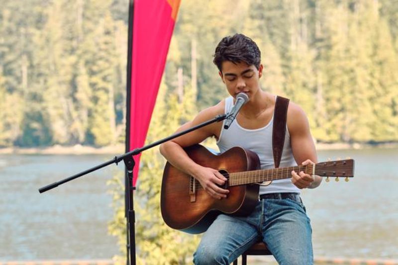 Un joven cantando concentrado frente a un micrófono mientras toca la guitarra acústica al aire libre con un lago y pinos de fondo.