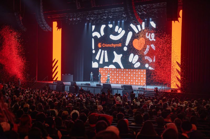 Presentación de Crunchyroll en Anime Expo 2026 Auditorio con público observando una presentación en escenario con elementos gráficos y paneles en tonos naranja y negro.