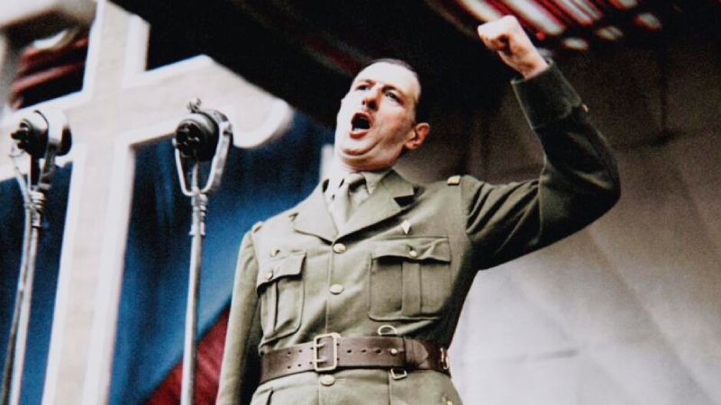 Segunda Guerra Mundial con Tom Hanks estrena episodios finales en HISTORY Fotografía histórica restaurada a color que muestra en plano medio al general francés Charles de Gaulle luciendo su uniforme militar color caqui y gorro militar. Se encuentra parado frente a dos micrófonos de radio clásicos y una gran estructura decorativa al fondo, alzando con vehemencia el puño derecho mientras exclama un discurso con la boca abierta durante un acto público.