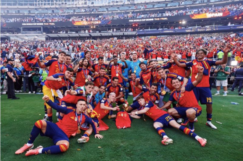 Seleccion Espanola celebra el titulo del Mundial 2026 docuserie Prime Video Fotografía deportiva en plano general en un estadio de fútbol iluminado con gradas llenas de aficionados. Al centro del campo de juego sobre el césped, los jugadores de la Selección Española celebran eufóricos vistiendo la camiseta roja oficial y medallas doradas al cuello; algunos posan acostados en el pasto, otros sentados y varios levantando las manos con expresiones de alegría junto a la réplica de la copa.
