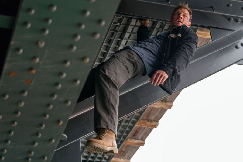 Alan Ritchson en escena de accion de la cuarta temporada de Reacher en Prime Video Fotografía de acción en plano medio con ángulo contrapicado. El actor Alan Ritchson interpretando a Jack Reacher cuelga de la estructura metálica inferior de un puente o viga industrial de acero con remaches. Viste una chamarra oscura con playera azul marino, pantalones cargo oscuros y botas de trabajo marrones; se aprecian raspones y heridas de combate en su cara y frente mientras mira hacia un lado con tono de urgencia.
