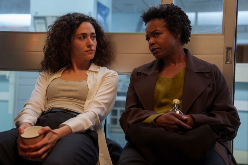 Emmy Rossum y Quincy Tyler Bernstine sentadas juntas en un pasillo conversando serenamente mientras sostienen vasos de café en un momento de pausa dramática.