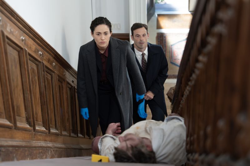 La agente Alice Black (Emmy Rossum) vistiendo abrigo gris y guantes de látex azules mientras sube cuidadosamente las escaleras de una residencia observando un cuerpo en el suelo durante una investigación policiaca.