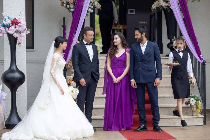 Escena de boda con personajes en conflicto Fotografía de una ceremonia con cinco personas en un entorno decorado con flores, telas y escalinatas, donde una pareja vestida de gala interactúa con otros invitados mientras una mesera desciende con una bandeja de bebidas.