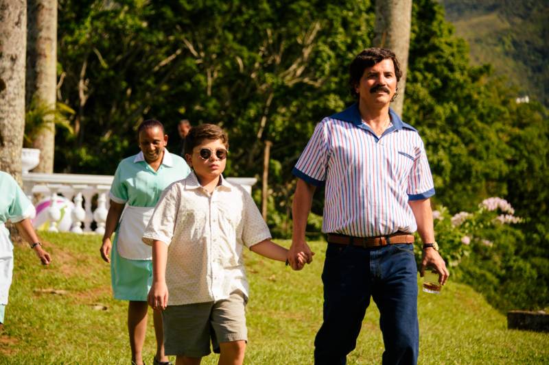 Escena familiar en entorno tropical Fotografía de un grupo de personas caminando en un área verde con árboles y montañas al fondo; un hombre toma de la mano a un niño mientras dos mujeres con uniformes claros caminan detrás.