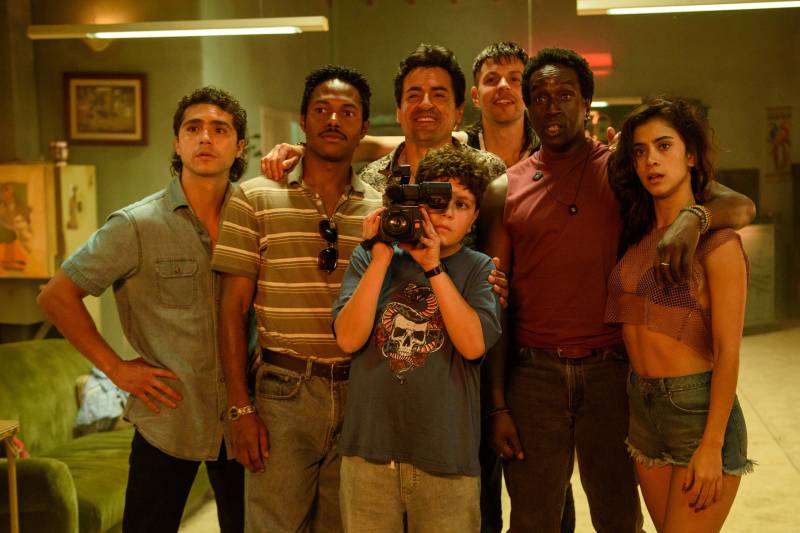 Elenco de Dear Killer Nannies en escena familiar Grupo de personajes reunidos en un espacio interior cálido, con un niño al centro sosteniendo una cámara mientras los adultos posan a su alrededor en un ambiente doméstico.