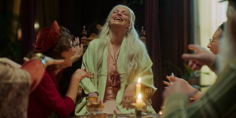 Un grupo de personas comparte una velada íntima alrededor de una mesa iluminada por velas, mientras una mujer mayor ríe con energía en el centro de la escena.