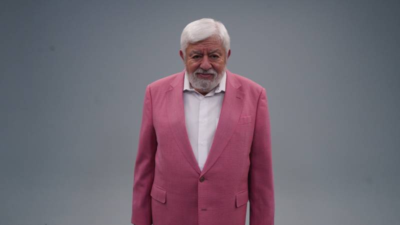 Persona de cabello blanco y barba posando con un saco rosa brillante frente a un fondo gris neutro, transmitiendo presencia y personalidad.
