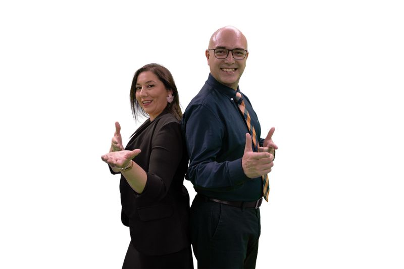 Una mujer vestida con saco negro y un hombre con camisa azul y corbata posan de espaldas juntos con las manos extendidas haciendo un gesto frente a un fondo negro.
