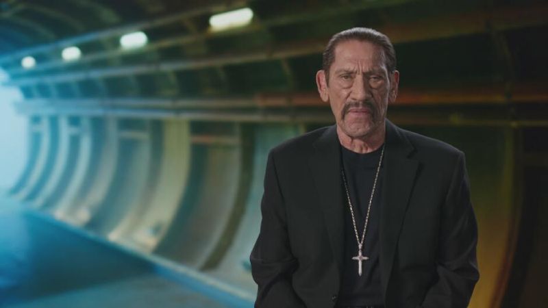 El actor Danny Trejo mira fijamente a la cámara en una toma de plano medio. Viste un saco negro, camiseta negra y un collar con un dije en forma de cruz plateada. Se encuentra posando en un estudio con un fondo abstracto de tonos azules que simula la estructura curva de un túnel o instalación industrial subterránea.