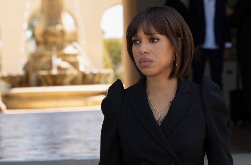 Retrato en plano medio de la actriz Kerry Washington interpretando a su personaje Eleanor. Luce un peinado estilo bob con fleco recto, viste un saco negro de vestir con escote cruzado y un collar dorado con un dije sutil. Mantiene una expresión seria y contemplativa mirando ligeramente fuera de cámara, con una fuente de agua y la entrada de una residencia iluminada al fondo en tonos cálidos fuera de foco.