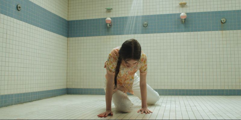 Escena de las Duchas en Carrie Summer H. Howell interpretando a Carrie arrodillada en el suelo de azulejos blancos de la regadera escolar bajo el chorro de agua.