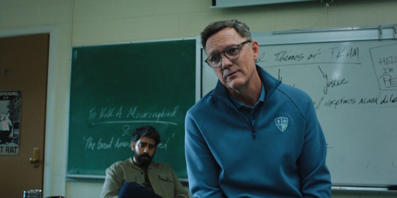 Docentes en el Entorno Escolar de Carrie Un profesor en primer plano hablando en el salón de clases con un pizarrón verde al fondo y un estudiante sentado detrás.