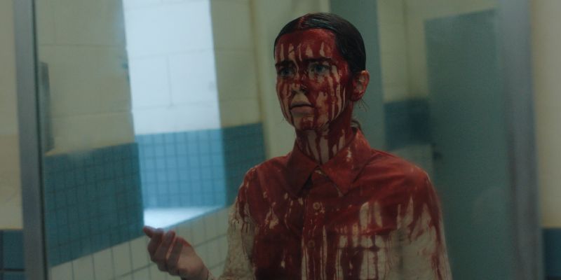 Carrie Bañada en Sangre frente al Espejo Carrie cubierta de sangre de pies a cabeza frente a un espejo mirando con una expresión de desconcierto y shock.
