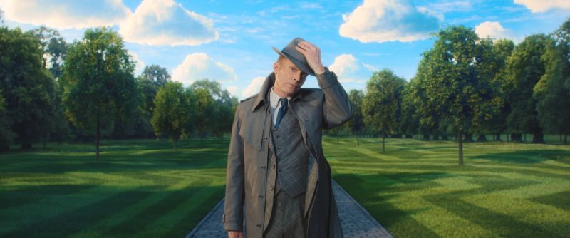 Paul Bettany como Visión en Su Espacio Mental Paul Bettany luciendo sombrero de ala y gabardina gris en un amplio jardín verde bajo un cielo azul con nubes.