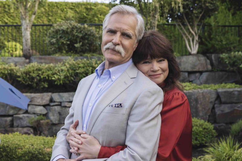 Los actores Henry Czerny y Valerie Bertinelli posan juntos al aire libre en un jardín rodeado de vegetación y muros de piedra. Henry Czerny, al frente, luce cabello canoso y un bigote blanco, viste un saco gris claro, camisa azul y un pañuelo de bolsillo estampado. Valerie Bertinelli lo abraza cariñosamente por la espalda rodeándole el pecho con los brazos, vistiendo una blusa roja de manga larga y sonriendo de forma serena hacia la cámara.