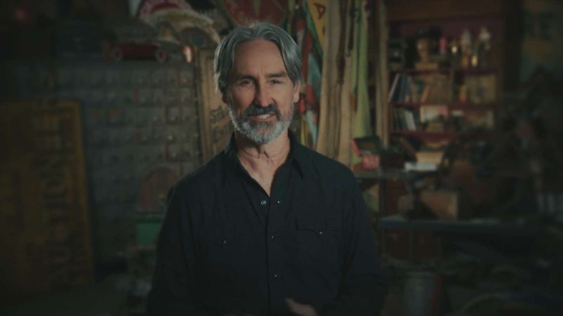 Mike Wolfe presenta Grandes Tesoros de la Historia en HISTORY El presentador Mike Wolfe mirando a la cámara con una sonrisa ligera. Viste una camisa de manga larga oscura estilo vaquero sobre un fondo de taller o depósito con herramientas, carteles vintage y estanterías de madera en tonos cálidos.