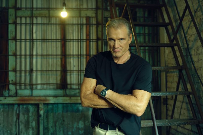 Dolph Lundgren presenta Grandes Maquinas de la Historia en History Retrato en plano medio del actor Dolph Lundgren vistiendo una playera negra y pantalón claro, parado con los brazos cruzados frente a una escalera metálica industrial dentro de una estructura de apariencia fabril con una bombilla encendida al fondo.