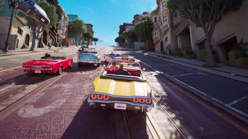 Carrera multijugador en Crazy Taxi World Tour de SEGA Captura de pantalla de jugabilidad de Crazy Taxi: World Tour. Muestra una amplia avenida inclinada al estilo de San Francisco en un día soleado, donde cuatro descapotables clásicos de colores amarillo, rojo y azul compiten a toda velocidad cuesta abajo. A la izquierda, un automóvil negro sale volando por los aires entre los árboles y edificios tras una colisión.