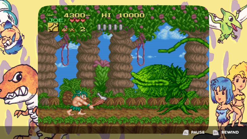 Gameplay de "Joe & Mac Retro Collection" con filtro CRT y marco ilustrado Captura de pantalla de jugabilidad (gameplay) en formato retro de "Joe & Mac Retro Collection". En el centro se observa a Joe en un escenario de selva prehistórica atacando a una planta carnívora gigante con un garrote de madera. La pantalla cuenta con un marco ilustrado con estilo de arte retro que incluye dinosaurios, cavernícolas y un pterodáctilo, además de mostrar los controles de pausas y rebobinado (rewind) en la barra inferior.
