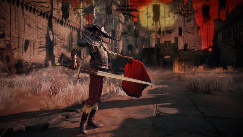 Captura de pantalla oficial de "ELDEN RING" centrada en un personaje ataviado con un conjunto de armadura estilizado con un sombrero de ala ancha y pluma, sosteniendo un mandoble y una capa o manto rojo contra un fondo de fortaleza medieval y un cielo rojizo.