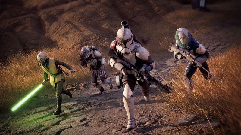Escuadrón de operadores en acción en Star Wars Zero Company Captura de pantalla de gameplay de Star Wars Zero Company que muestra a un escuadrón de cuatro operadores avanzando en fila por un camino de piedra en un entorno árido con hierba seca. En primer plano, al centro, encabeza la Marcha un Clon Trooper con armadura blanca desgastada y marcas de batalla. A la izquierda, una joven Padawan alienígena vistiendo túnicas Jedi sostiene un sable de luz de hoja verde encendida. Detrás del Clon avanza un guerrero Mandalorian con armadura blanca y roja equipada con jetpack. A la derecha, un tirador de élite porta un casco protector de burbuja transparente y un rifle de precisión.