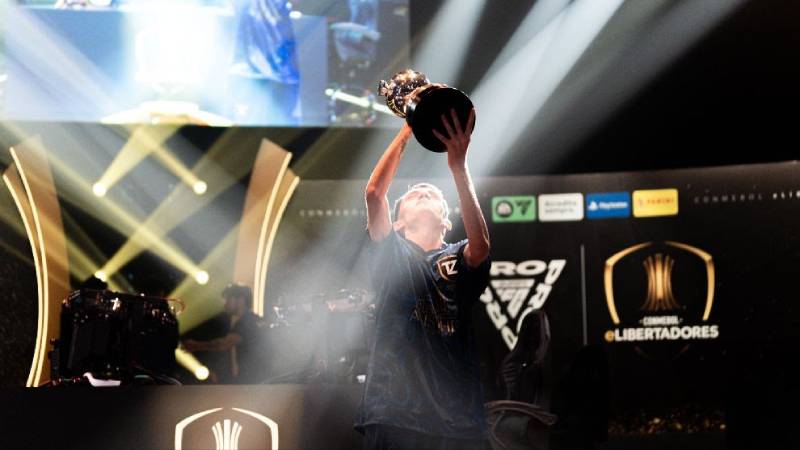 Celebración en torneo de esports de CONMEBOL eLibertadores Fotografía de un jugador levantando un trofeo en un escenario iluminado, rodeado de pantallas y elementos visuales propios de un torneo profesional de esports.
