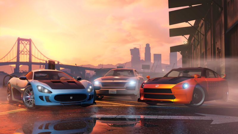 Escena urbana con autos deportivos en “GTA Online” Tres autos deportivos estacionados sobre una calle mojada al atardecer, con un puente y edificios urbanos al fondo.