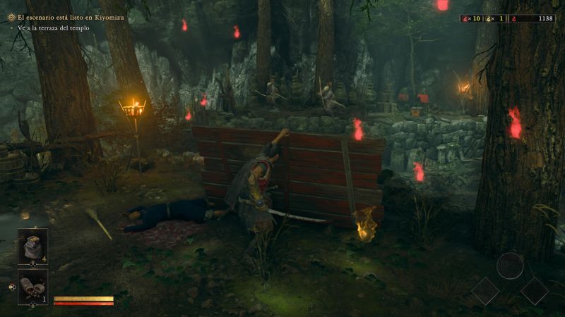 Mecánicas de acción e interacción con el entorno Musashi agazapado detrás de una barricada de madera en un bosque oscuro iluminado por antorchas y almas rojas flotantes, acechando a enemigos a lo lejos.